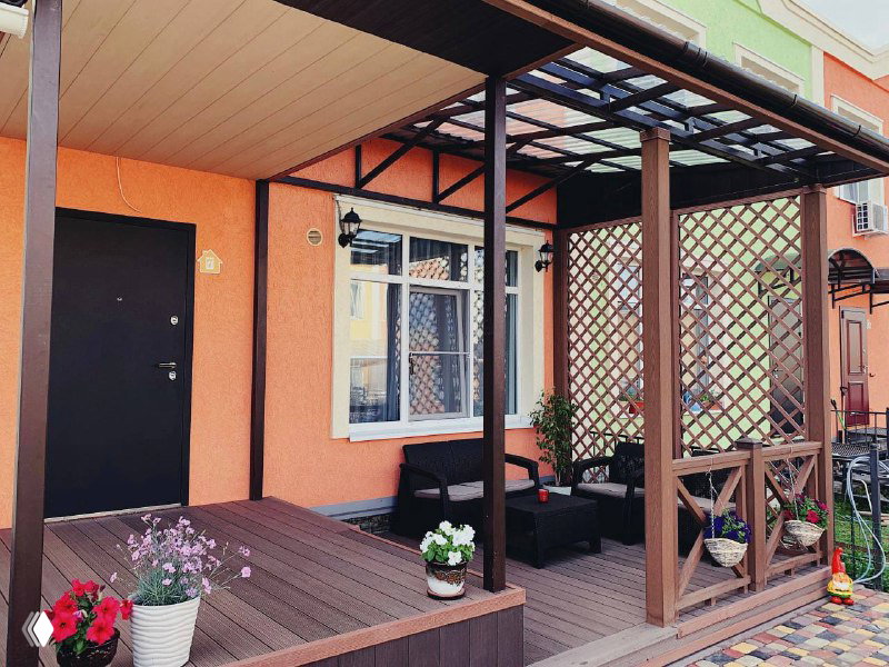 Маленькая веранда перед домом с настилом из ДПК‑доски, цветочные горшки и мебель на фоне фасада, показано применение ProDecking в частном доме.