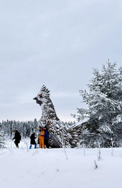 Высокая деревянная птица‑рябчик, собранная из брусочков по технике Вальковой; рядом люди у подножия, зимний лес и снег вокруг создают контекст.