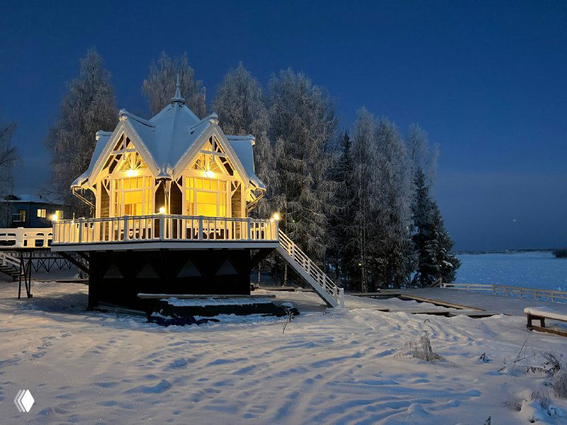 Небольшой домик-музей в Голубино у берега в зимний сумрак; тёпло освещённый домик на сваях, вокруг заснеженные деревья и замёрзшая вода.