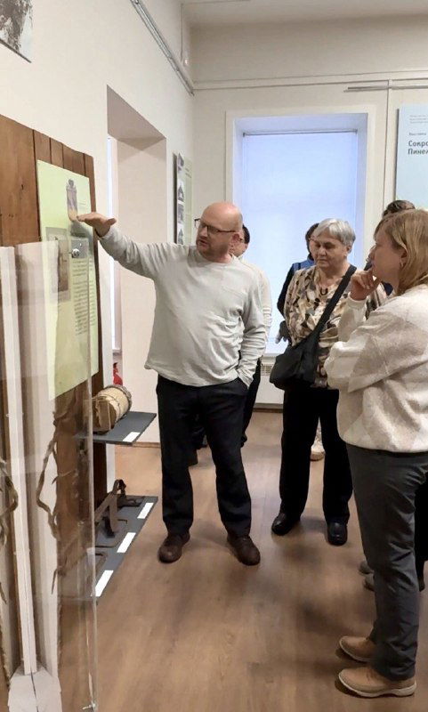Экскурсовод Константин рассказывает группе посетителей у музейной витрины, видна часть аудитории и экспонатов на стене.