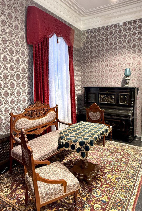 Ещё один интерьерный зал с салоном XIX века: диван, стол и пианино на фоне обоев и ковра, воссоздана домашняя обстановка.