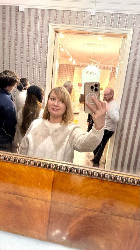 Автопортрет посетительницы в зеркале музейной комнаты: отражение группы гостей и часть интерьера на заднем плане.