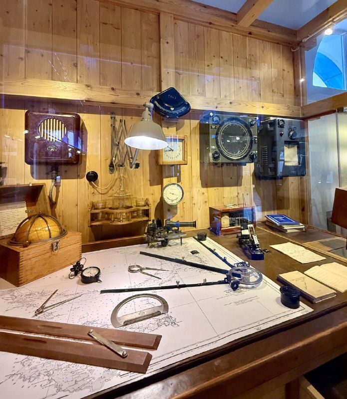 Рабочий стол с навигационными приборами: циркули, секстант, чертежи и инструменты на деревянной панели в музейной экспозиции по судоходству.