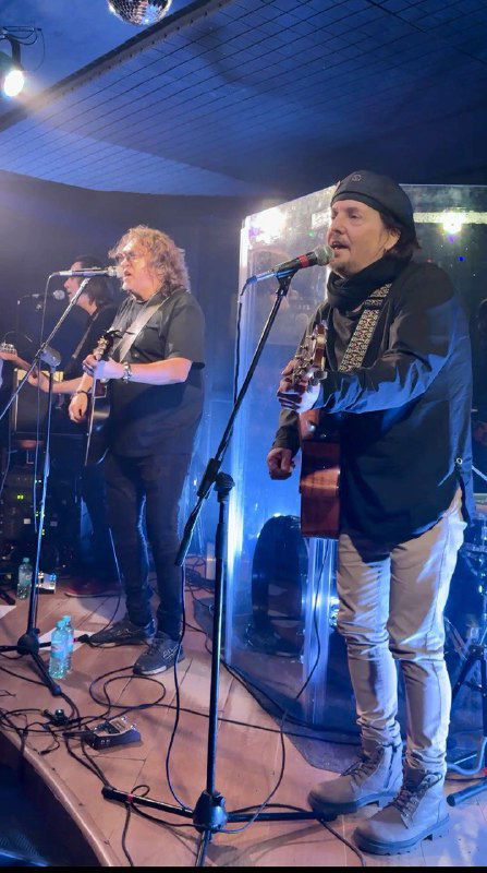 Группа на сцене клуба «Колесо»: два музыканта поют и играют на гитарах при сценическом освещении во время концерта