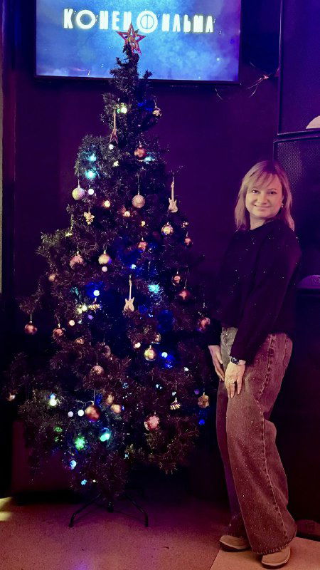 Девушка стоит рядом с украшенной ёлкой в интерьере клуба «Колесо», праздничные огни и декор создают новогоднее настроение
