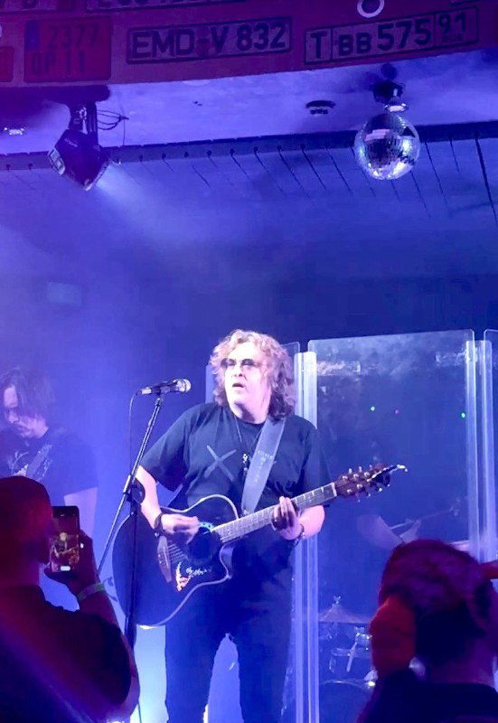 Гитарист на сцене во время концерта в небольшом музыкальном клубе, видны зрители и тёмный интерьер зала с подсветкой