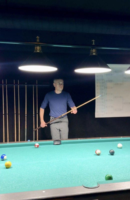 Мужчина играет в американский пул в бильярдной: стол с шарами, зелёное сукно и лампы над столом, уютно и по-домашнему