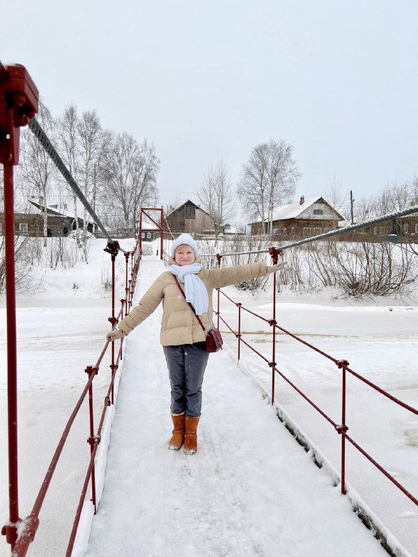 Женщина идёт по подвесному пешеходному мосту зимой в тёплой одежде, вокруг заснеженные деревни и деревянные дома на берегу реки