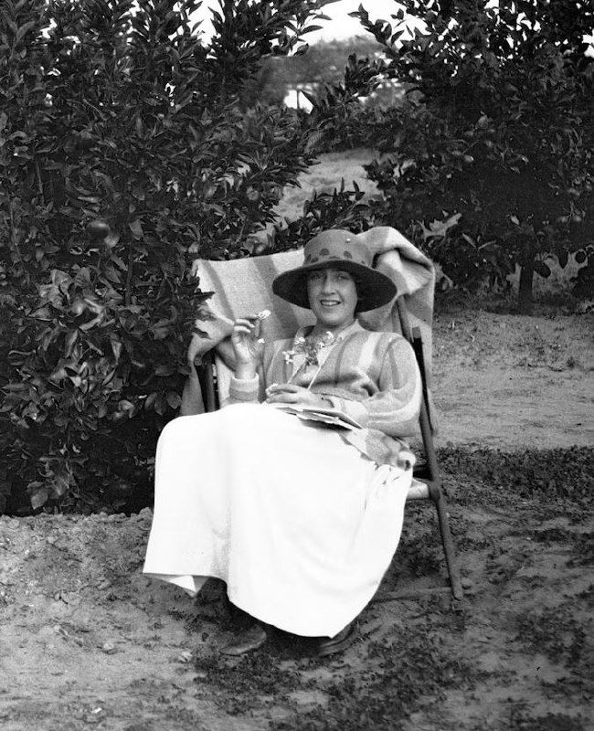 Черно-белое фото Агаты Кристи в летней шляпе, отдыхающей в кресле на природе; рядом чайник и стакан, мягкая улыбка на лице.