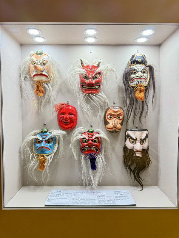 Коллекция театральных масок разных стилей и выражений, аккуратно размещённых в витрине — часть раздела о масочной традиции.