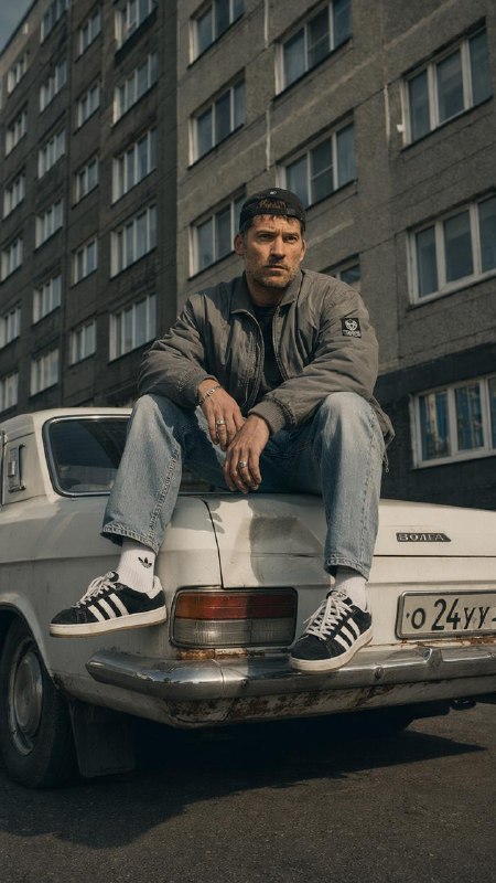 Мужчина в бомбер‑куртке и кедах сидит на багажнике Волги; в кадре ритмичная фасадная стена дома, следы ржавчины и городская фактура асфальта.