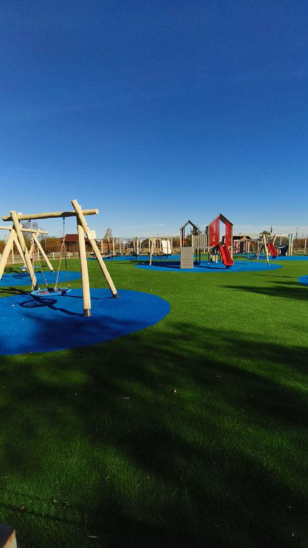 Яркая детская площадка с качелями и игровыми конструкциями на мягком покрытии в зелёной зоне базы отдыха под чистым небом.