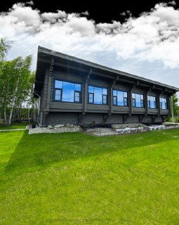 Длинное тёмное здание апартаментов или коттеджей на фоне газона и неба, аккуратный ландшафт вокруг комплекса.