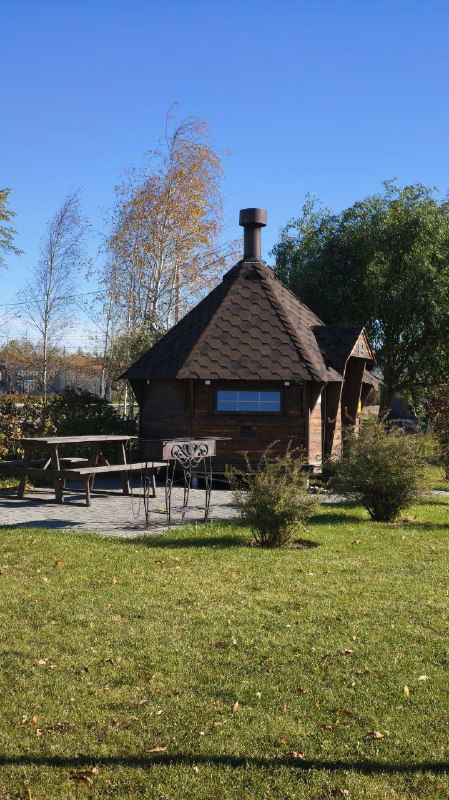 Небольшая деревянная баня с зоной для барбекю и столиками на газоне, зелёная территория вокруг и осенние деревья.