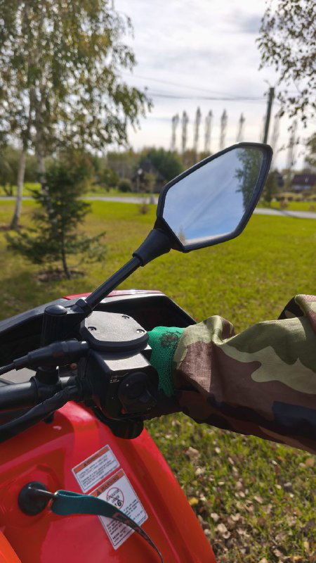 Крупный план левого зеркала и руля квадроцикла, видна рука в защитной перчатке, на заднем плане парк и промышленные сооружения.