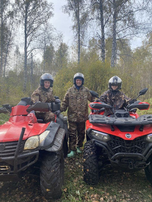Трое участников в камуфляже стоят между двумя красными квадроциклами в лесной полосе, видны берёзовые деревья и подготовка к маршруту.