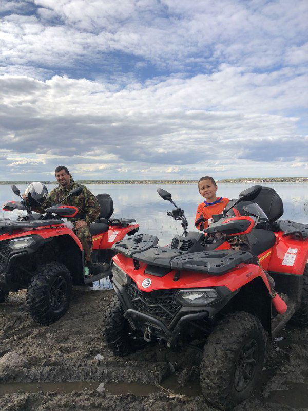 Два человека на красных квадроциклах на прибрежной грязной дороге, на переднем плане шлем и брызги грязи, солнечное небо над озером.