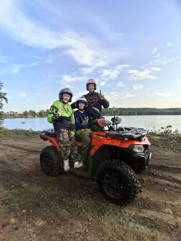 Два ребёнка в шлемах и взрослый на квадроцикле на фоне озера, улыбки и дружелюбная атмосфера семейного отдыха на свежем воздухе.