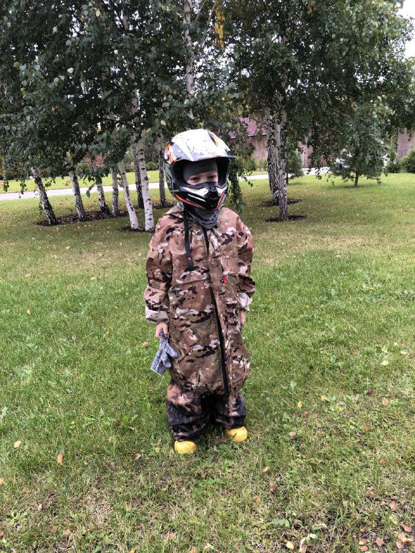 Маленький ребёнок в камуфляжном костюме и шлеме стоит на траве возле берёз, подготовка к выезду и детская экипировка в естественной обстановке.