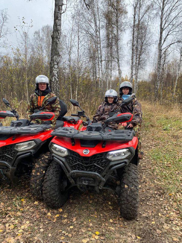 Три ребёнка в шлемах сидят на квадроциклах в лесу, видны лиственные деревья и дождливо-осенняя погода, безопасная организация детского катания.