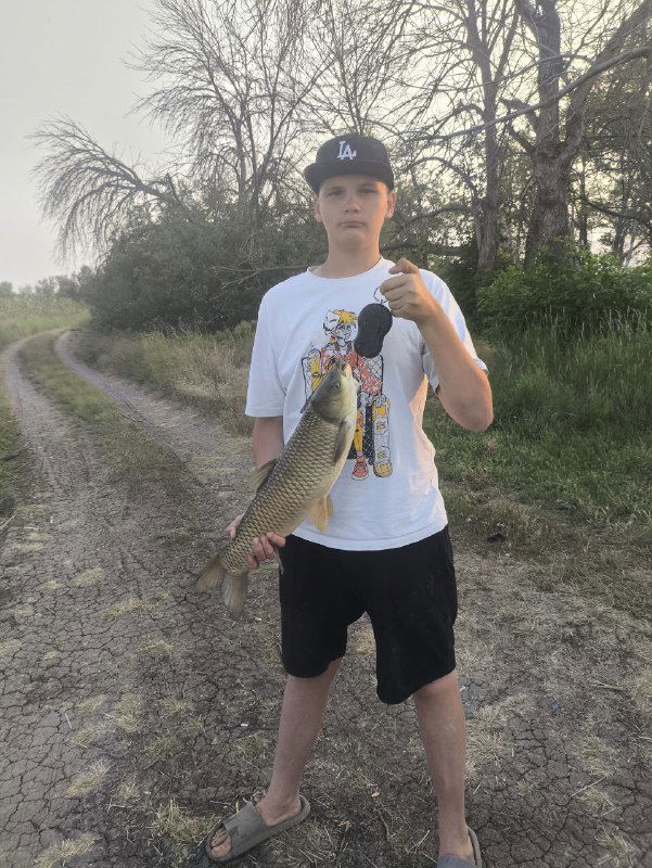 Рыбак стоит на грунтовой дороге у воды и держит крупного амура на вытянутых руках; фон — деревья и летний пейзаж донских берегов.