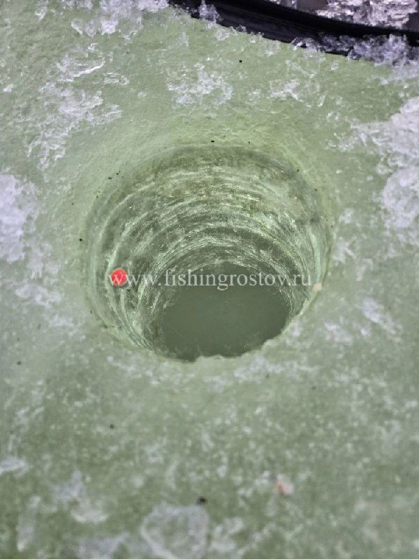 Крупный план просверленной лунки во льду с заметной толщиной и зелёным оттенком на поверхности — видна вода в отверстии.