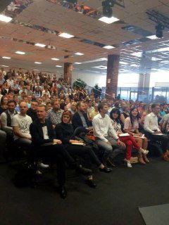 Фотография большой аудитории конференции: заполненный зал участников, люди сидят в рядах, видна сцена и общий масштаб мероприятия.