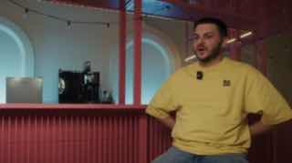 Кадр из интервью: сотрудник в жёлтой толстовке сидит в студии перед красной стойкой — фрагмент видео о биздеве и продажах, иллюстрация выпуска