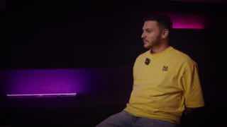 Кадр интервью: мужчина в жёлтой футболке сидит в студии с тёмным фоном и пурпурной подсветкой, разговорная сцена.