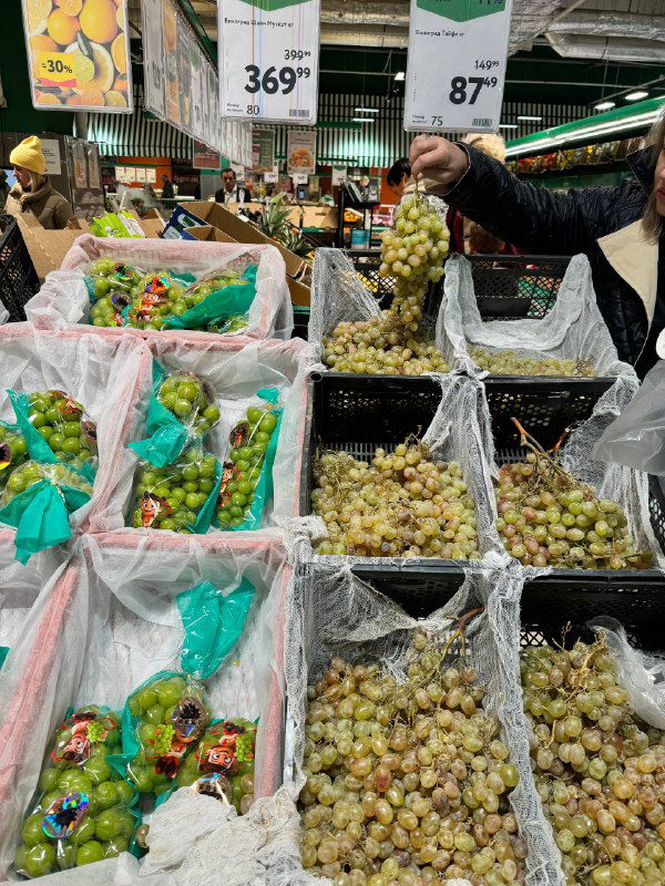 Ряды винограда в лотках магазина Окей: разные сорта в пакетах и открытые корзины, покупатель выбирает гроздь у прилавка.