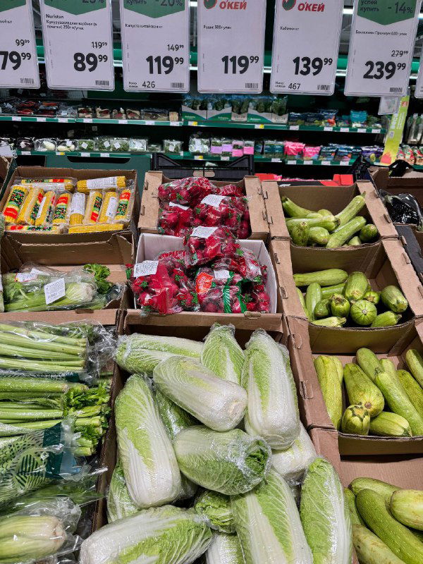 Ассортимент зелёных овощей и упаковок с белокочанной и китайской капустой на прилавке с ценниками в супермаркете Окей.