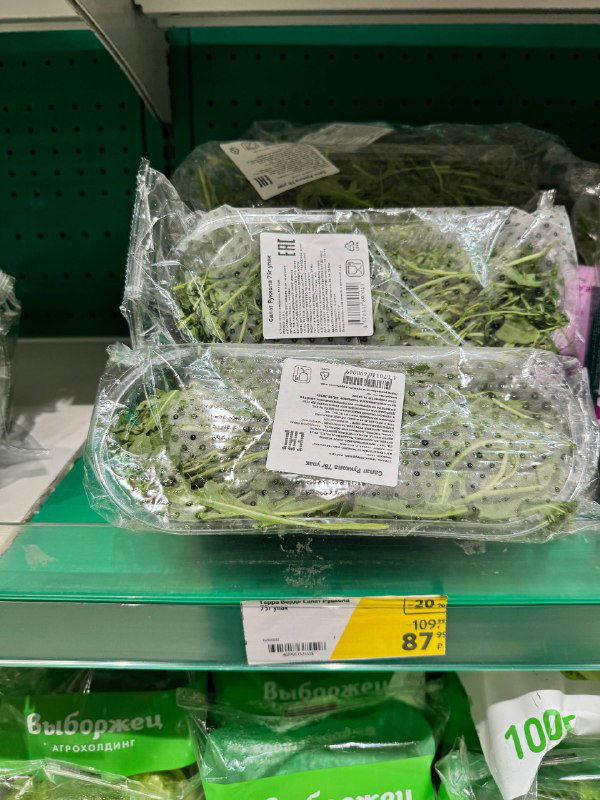 Упакованная зелень на полке супермаркета: руккола, шпинат и другие листовые культуры с промо-ценами и ярлыками.