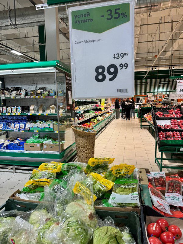 Промо-стойка с пакетированной зеленью и акционной ценой, общий вид торгового зала и прилавков с овощами.