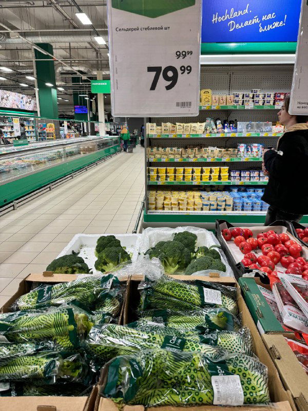 Лоток с брокколи и другими зелёными овощами, ценник 79–99₽ виден над прилавком, супермаркет в рабочее время.