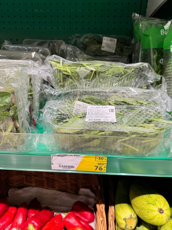 Пакетированная зелень и травы на полке супермаркета, видны штрих-коды и информационные стикеры на упаковках.