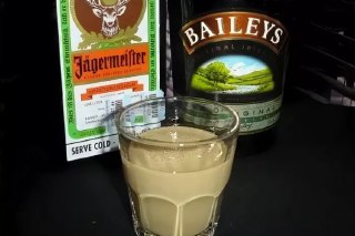 Стаканчик с кремовым бежевым коктейлем на фоне бутылок Jägermeister и Baileys — визуал для рецепта «Бешеный пёс».