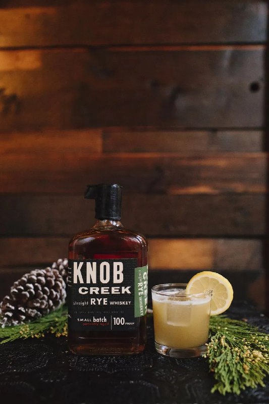 Бутылка Knob Creek рядом с маленьким стаканом коктейля на деревянном столе и еловыми ветками — атмосферный зимний кадр.