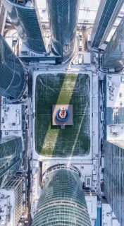 Аэрофотосъёмка: городская спортивная площадка между небоскрёбами, в центре — батут и маленькая фигура человека, кадр сверху.