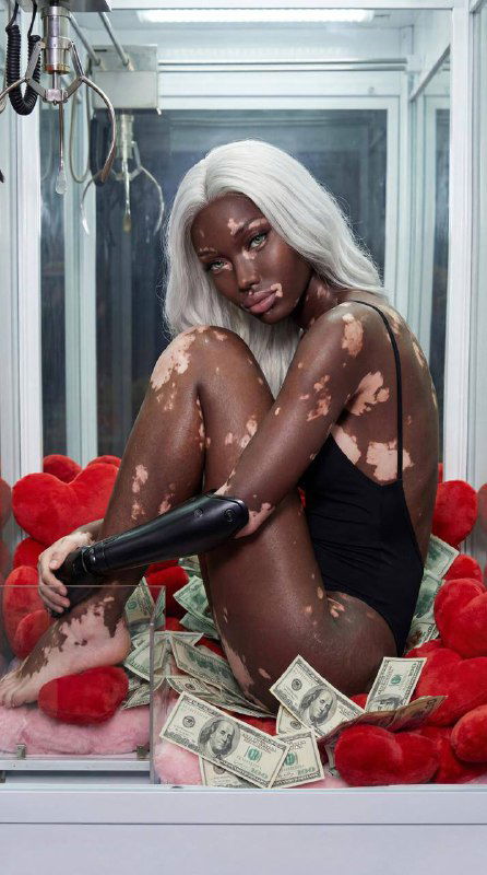 Портретная композиция: модель с контрастной кожей и светлыми волосами сидит в автомате, вокруг мягкие красные подушки в форме сердец и купюры на фоне стекла.