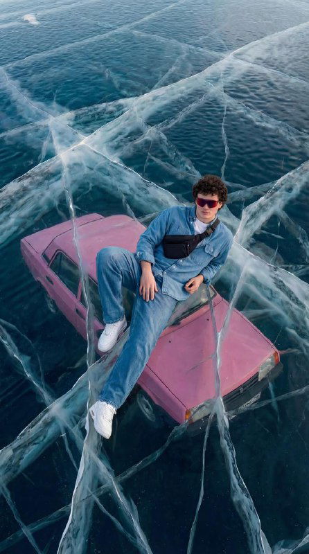 Вид сверху: мужчина в джинсах и кроссовках сидит на розовом автомобиле, лёд с крупными трещинами и прозрачной структурой.
