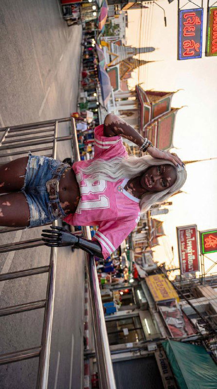 Широкий стрит-кадр: модель в джинсовых шортах и розовой футболке на городской улице с торговыми палатками и неоновыми вывесками на фоне.