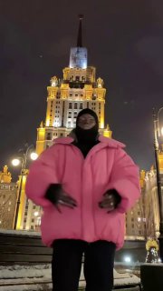 Крупный вертикальный план человека в розовой куртке и капюшоне на ночной площади перед освещённым архитектурным фасадом и уличными фонарями.