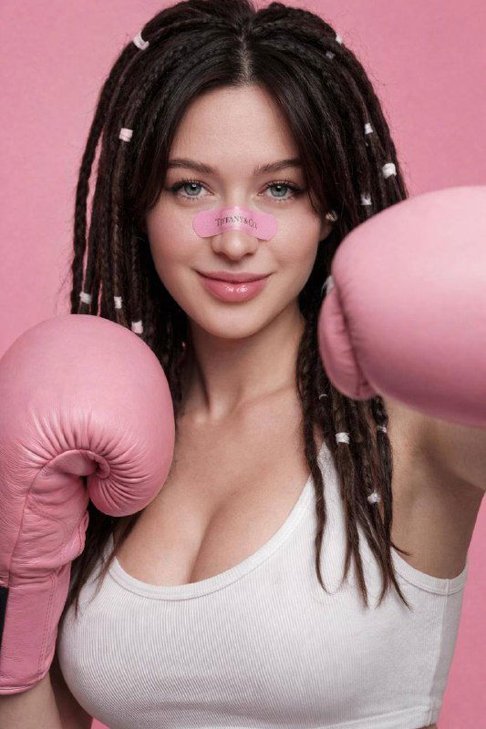 Крупный план молодой женщины с дредами, белая майка и розовые боксерские перчатки на однотонном фоне, пластырь на переносице.