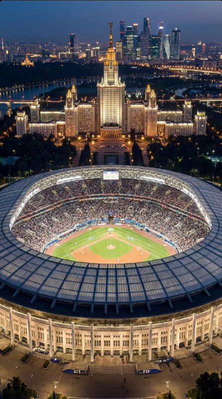 Ночной панорамный вид на освещённый бейсбольный стадион и городской пейзаж вдалеке, вид сверху с акцентом на арену и огни.