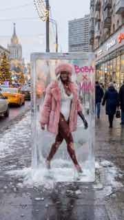 Уличный вертикальный снимок: модель в розовом образе внутри ледяного блока на городском тротуаре зимой, прохожие и мокрый асфальт.