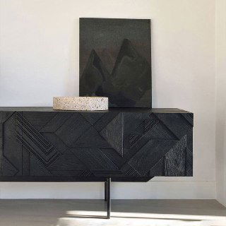 Крупный план фасада тумбы из ясеня с геометрическим резным узором и матовой чёрной отделкой на фоне светлой стены в комнате.