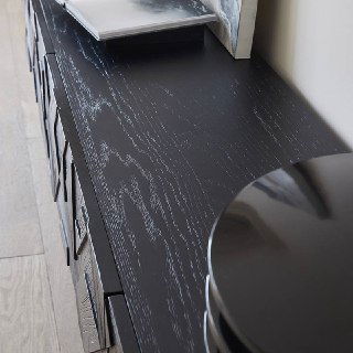 Верхняя поверхность тумбы из ясеня с видимыми волокнами древесины и матово-чёрной отделкой, предметы декора частично видны на столешнице.