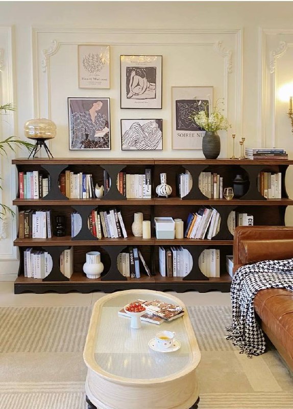 Широкая низкая книжная полка из ясеня с множеством горизонтальных отделений, декорирована вазами и книгами, вписанная в светлый интерьер гостиной.