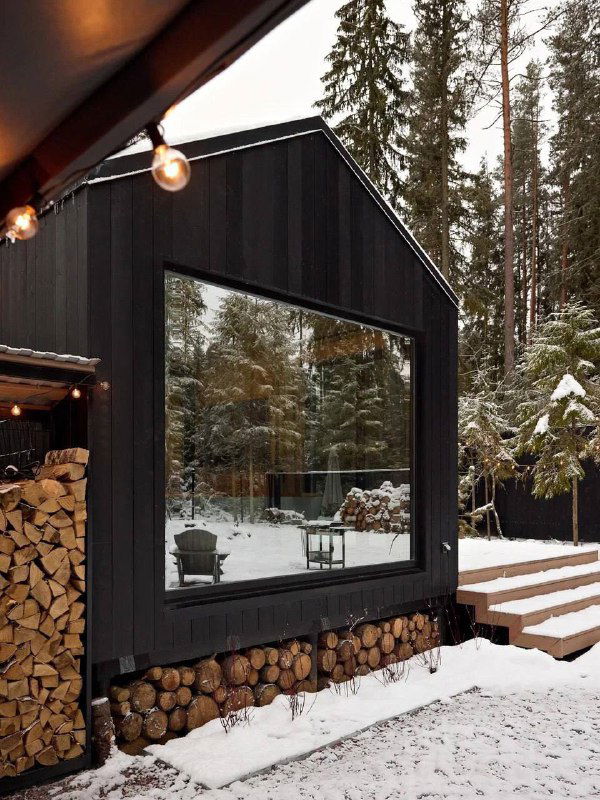 Крупный план большого панорамного окна в деревянном фасаде, отражение леса в стекле и аккуратно сложенные поленья под окном, снег вокруг.