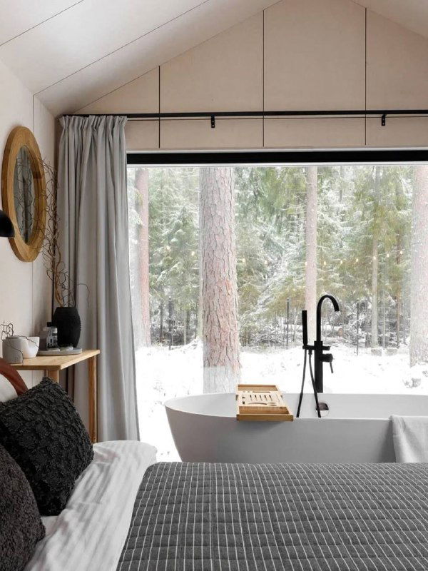 Спальня с окном от пола до потолка, отдельная ванна у окна и вид на заснеженные деревья — сочетание уюта и природы для отдыха.